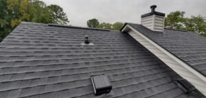 Overhead view of a gray shingle roof with vents and a chimney, with trees in the background under a cloudy sky