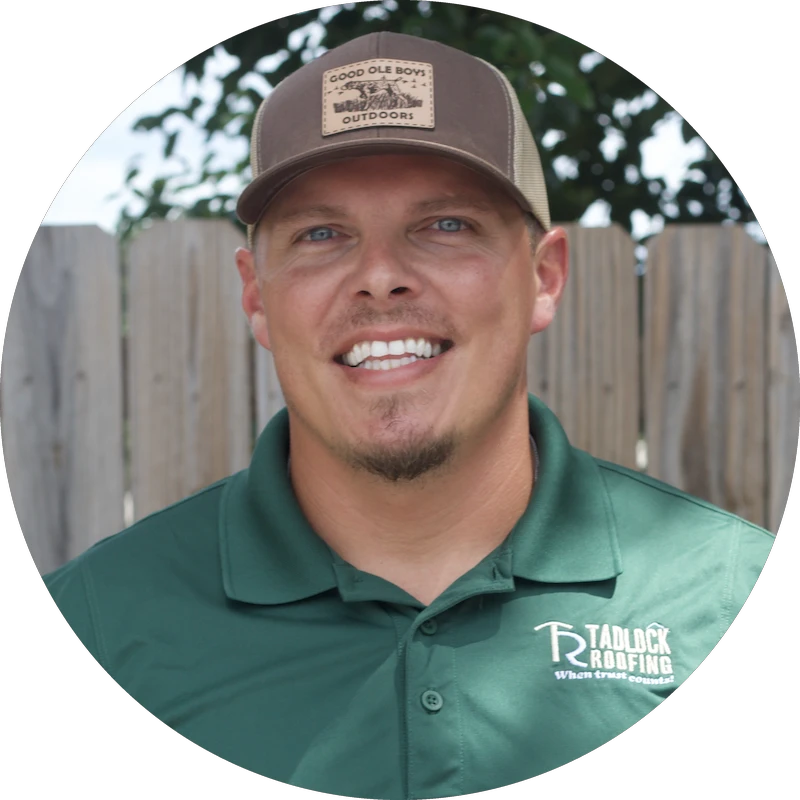 Smiling man in a Tadlock Roofing green polo and brown cap, standing outdoors by a wooden fence.